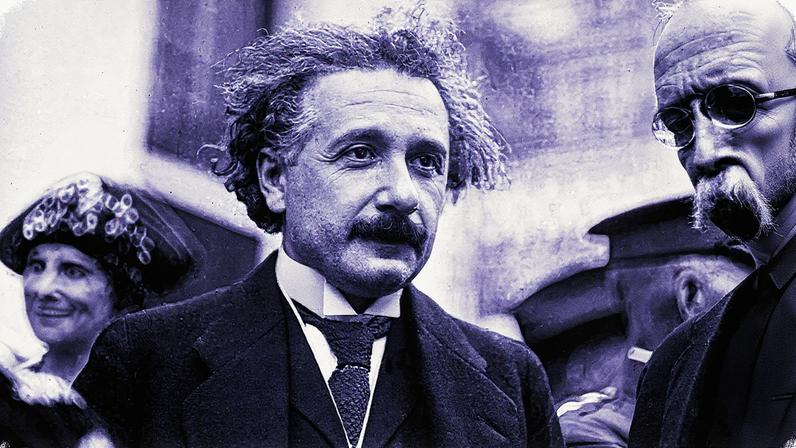 Homem com cabelo bagunçado e bigode, usando um paletó escuro, sentado em uma mesa com papéis e um livro aberto (Foto: Reprodução)