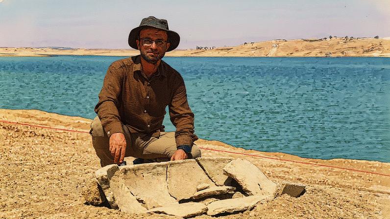 Bekas Brefkany, diretor do Departamento de Antiguidades de Dohuk, posa ao lado de uma sepultura descoberta nas margens da Barragem de Mosul em um sítio arqueológico no subdistrito de Khanke, em Dohuk (Foto: Reprodução)