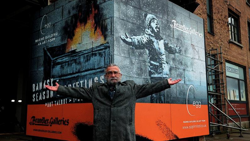 Homem de terno em frente à obra "Season's Greetings" de Banksy em 2022 (Foto: Reprodução)