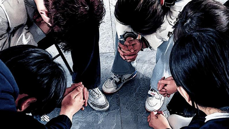 Um jovem é visto da perspectiva de cima. Ele tem cabelos curtos, usa óculos e tem duas mãos impostas em seus ombros em oração. No texto lê-se Ajude a treinar líderes de jovens na China